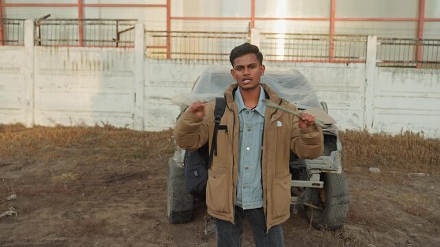 Standing by wrecked car showing parts, young man raises broken wipers then approaches camera in open salvage yard wide shot near fence with jacket and backpack, confident presentation and DIY repair