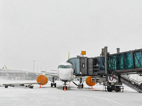 A commercial airplane is stationed at a snowy airport, connected to a jet bridge. The aircraft is surrounded by snow-covered tarmac, with several orange engine covers visible.
