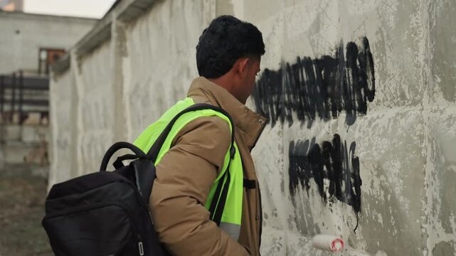 Painter rolling paint over graffiti on concrete wall, reflective vest and backpack visible, brisk brush and roller strokes cover vandal marks, focused city repair and cleanup action at jobsite