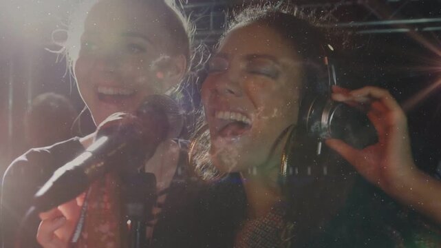 Two women walking into frame, gathering at mic, cupping headset, harmonizing music under flare haze