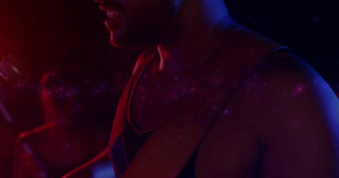 Singing man wearing tank top, guitar strap, leaning into mic starting song at small club, sparkles