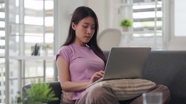 Asian adult woman working from home online using laptop computer on comfortable couch productivity freelance business education virtual