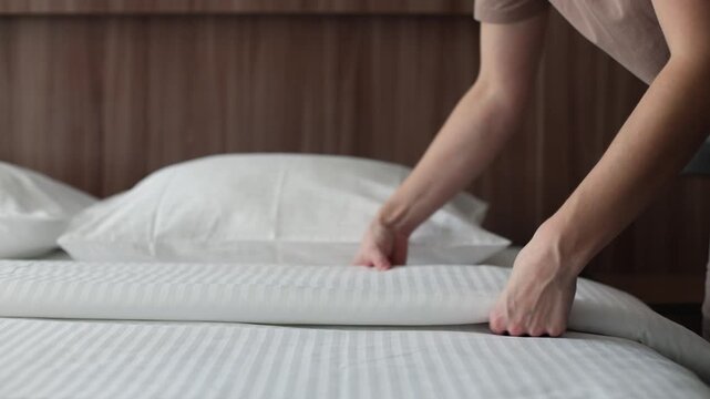 A maid in a hotel room makes the bed, smoothing out the duvet with her hands.