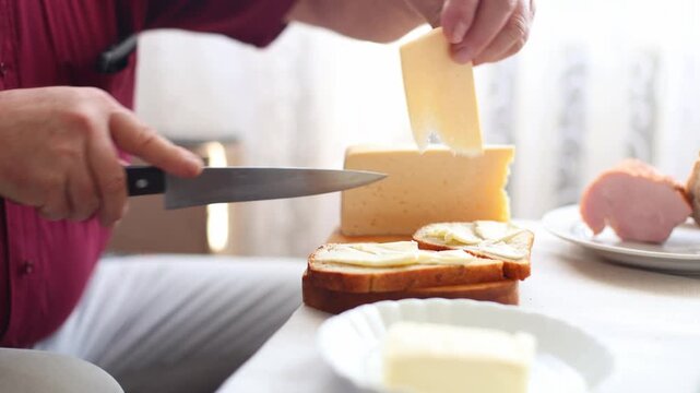 Close-up of hands slicing yellow cheese with a knife and placing it on buttered bread, breakfast prep in a bright, homey setting.