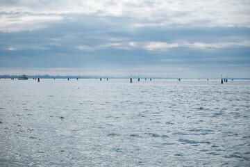 Obraz premium Venice, Italy - February 10, 2026: Calm water surface with wooden poles emerging, cloudy sky above and distant boat on the horizon in a coastal setting