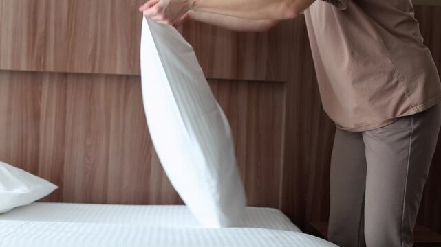 A hotel staff member fluffs and arranges white pillows on a bed with a wooden headboard in a modern room.