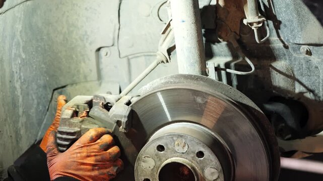 A close-up video shows a mechanic wearing orange gloves servicing a car&rsquo;s front brake system, carefully positioning the brake caliper over the disc and applying lubricant to ensure smooth 