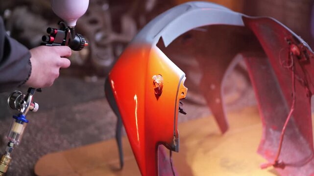 A close-up video captures a mechanic spray painting a motorcycle plastic fairing, applying a glossy orange-red coat over a primed surface using a professional spray gun with pressure regulator, 2026