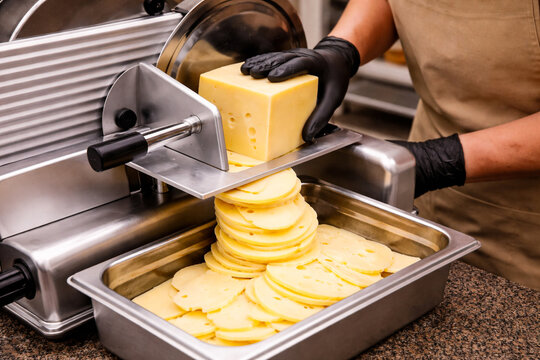 Close-up of a worker wearing black gloves slicing a block of cheese on an electric slicer in a deli or supermarket. Thin slices fall into a tray, showcasing food preparation and retail process.
