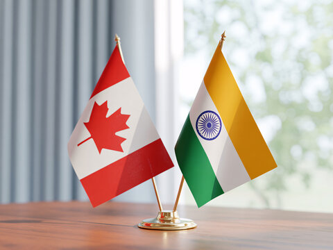 The flags of Canada and India stand side by side on a wooden table in a bright room.
