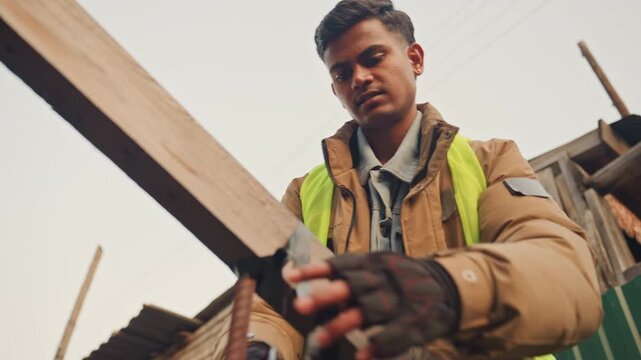 Worker measuring wooden beam for repair and alignment. Asian construction worker in highvisibility vest and gloves uses tape and tools to check timber join, secures hardware and prepares framework