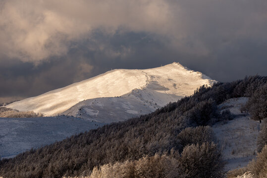 Dramatic winter view of Smerek peak in Bieszczady Mountains illuminated by golden sunlight against dark stormy clouds.