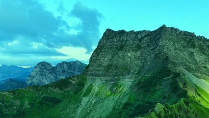 Fotobehang Turkoois Aerial view of alpine mountain cliff with dramatic sky and textured rock layers, cinematic landscape with green slopes and natural patterns under soft evening light  © Drones Sauvages