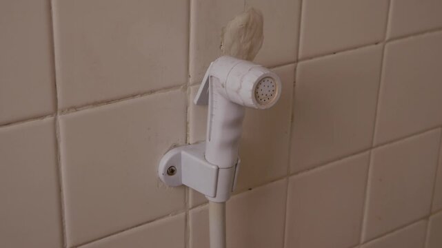 White plastic bidet hand shower sprayer mounted on clean beige square ceramic tile bathroom wall with white hose