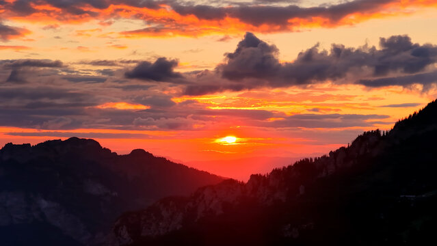 Aerial view of dramatic sunset over alpine mountains with glowing sky, vibrant orange clouds and silhouetted peaks creating a cinematic landscape
