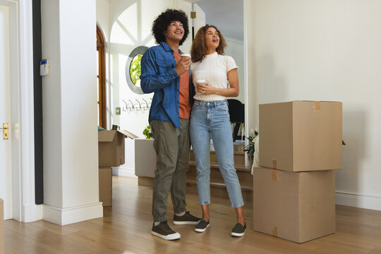Diverse couple standing, holding takeaway coffee cups, looking upward in entryway near boxes