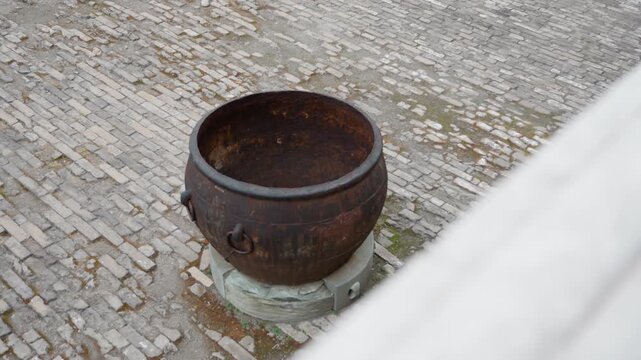 Rusted iron cauldron on cobblestone courtyard, weathered pedestal and peeling patina evoke historic decay overhead perspective shows textured paving, subtle moss growth, muted tones and moody