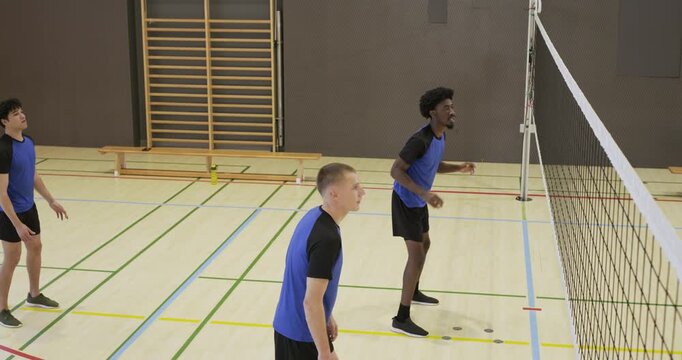 Three men in blue jerseys jumping, driving ball at net in gym, winning point, forming triple-block