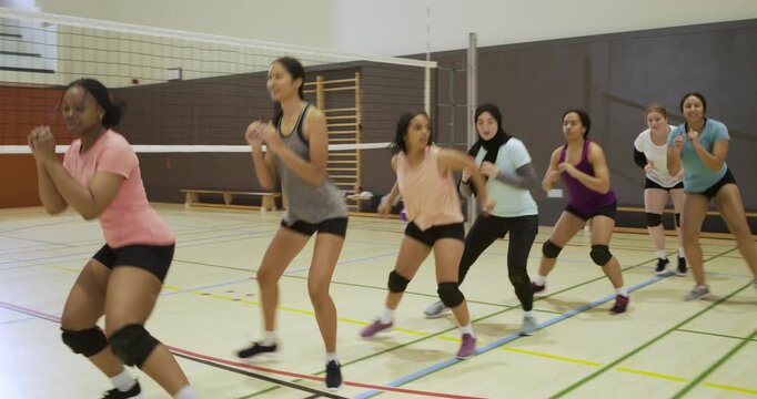 Female volleyball team performing high-knee hops warming up on gym court with coach signaling, net