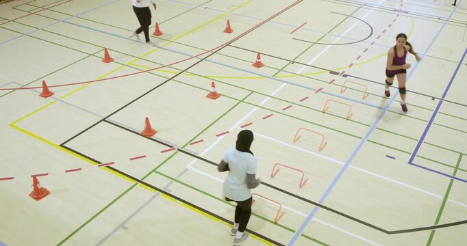 Female teammates in sportswear following coach signal, running cones and hurdles toward hoop in gym