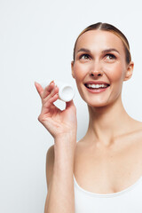 Young smiling woman with clear skin holds white cosmetic jar near face looks up happily isolated on...