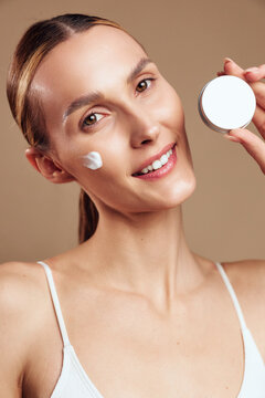 Young woman applying cream on face smiling looking at camera natural skincare routine healthy skin soft lighting beige background beauty concept