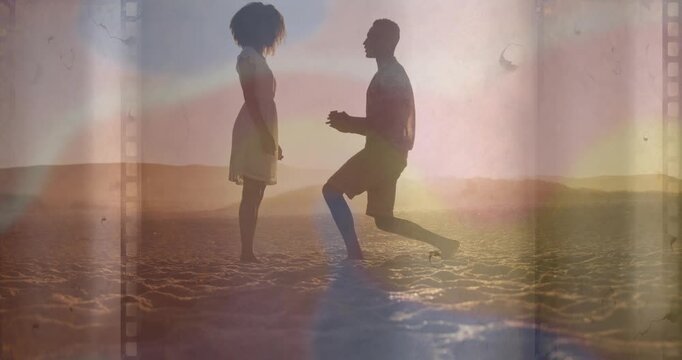 Man walking and kneeling on sand proposing with ring while she reaching film overlay framing couple