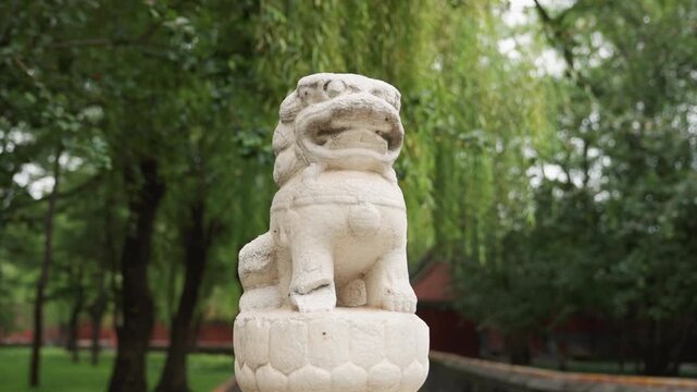 Stone guardian lion statue in willow park, textured limestone carving on pedestal, tranquil courtyard setting, gardener tending nearby beds, soft overcast light, moss accents, heritage atmosphere,