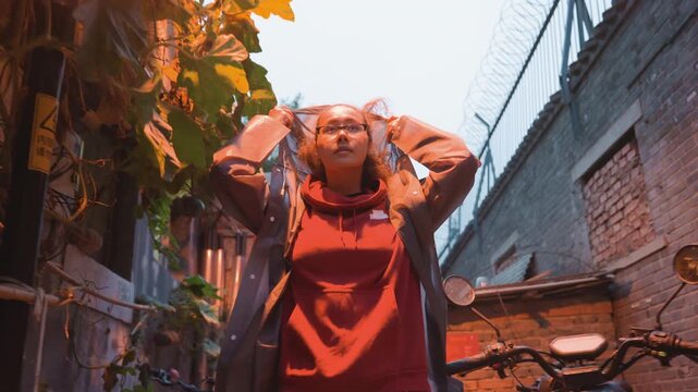 Asian woman walking narrow alley dusk student removing hood, curious gaze to camera, red hoodie under gray coat, brick walls and vine foliage, warm streetlamp glow, bicycle and local signage, intimate