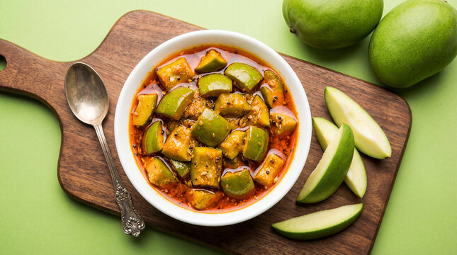 A bowl of spicy raw mango pickle or aam ka achar served with fresh green mangoes on a wooden board