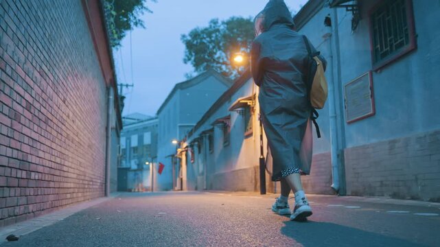 Female explores alleyway. Female Asian individual moving rapidly through city passageway with backpack. An Asian woman briskly proceeding down alley with yellow backpack and lamplight glare