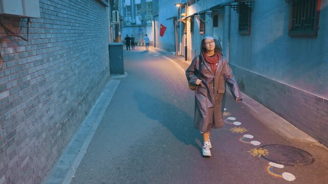 Smiling woman striding down lit alley wearing long coat and red scarf with sneakers on painted pavement, making eye contact with camera, upbeat confident walk, warm streetlights and brick walls,