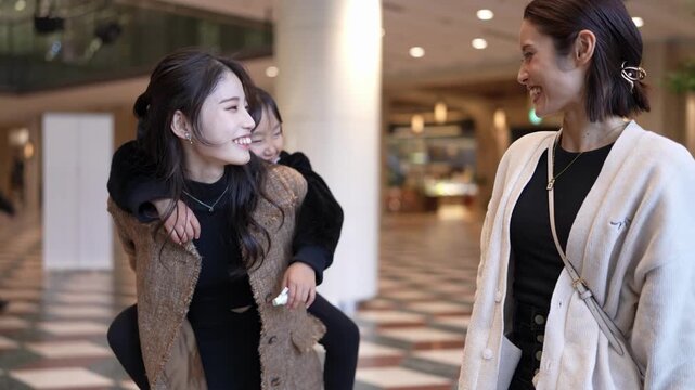 Two women share laughter in a well-lit mall corridor. One woman hugs another, both smiling warmly. Third woman joins them, enjoying the cheerful moment. The setting is casual and relaxed