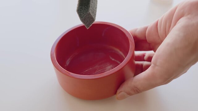 Hand Holding Red Bowl While Applying Paint With Foam Brush in Closeup Crafting Scene