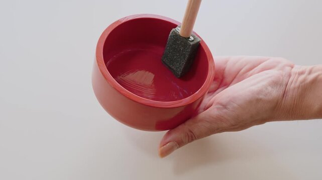 Hand Holding Red Bowl While Applying Paint With Foam Brush in Closeup Crafting Scene