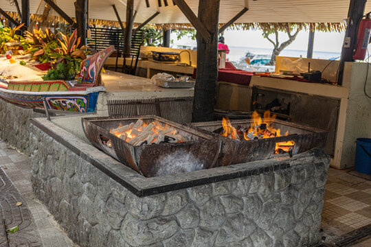 Charcoal grill fire pits blazing in a seaside tropical restaurant, next to a colorful decorated boat and fresh seafood display on ice, coastal dining atmosphere for tourists.