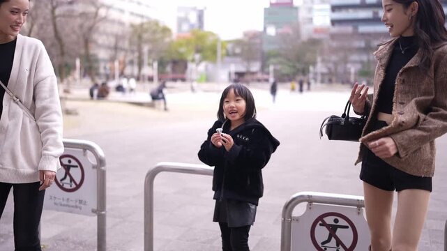Three women and a child stroll through a city park. One woman gestures wide, smiling joyfully. Another woman laughs, clapping her hands. The child stands in the center, arms wide open