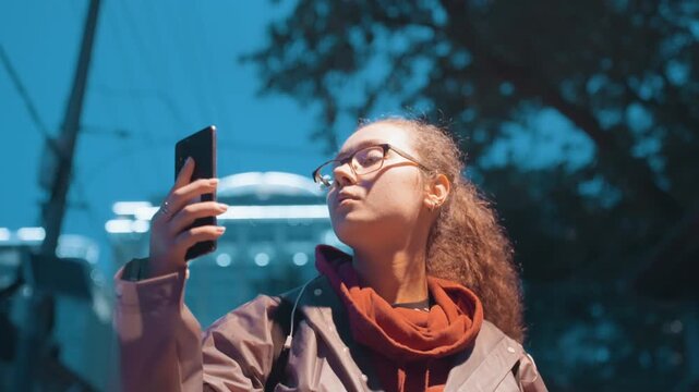 Glasses woman checking phone city lights, scarf wrapped against cool air, focused upward gaze at streetlamp glow, tapping screen for directions, reflective windows and neon backdrop, candid navigation