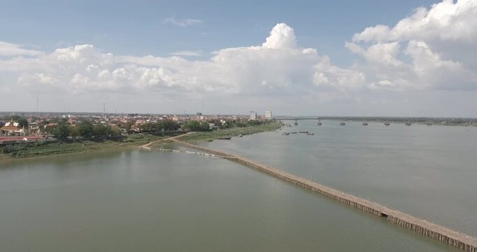 Wide aerial view of the long bamboo bridge crossing the Mekong River at Kampong Cham, revealing the city skyline and a modern bridge under a cloudy sky.