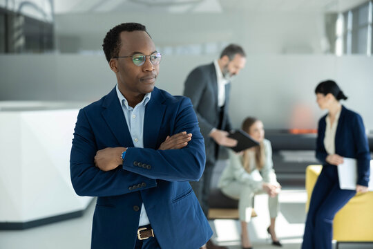 Young african entrepreneur with crossed arms posing confidently in a corporate setting with diverse colleagues in the background