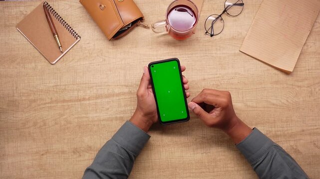 Man using smartphone with green screen on wooden table with tea and notepad.