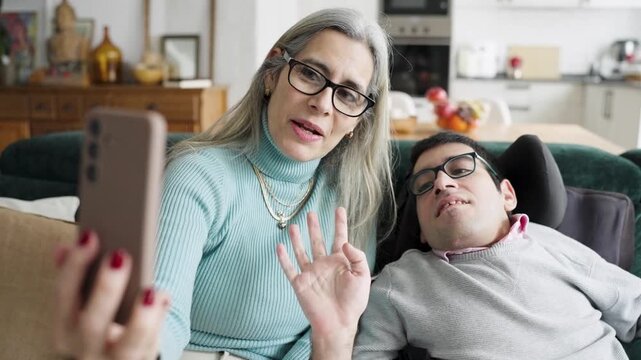 Happy mother and son with a physical condition sitting on the sofa at home, smiling and waving during a video call on a smartphone
