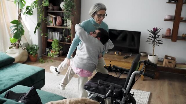 Loving mother carefully lifts her son with a physical impairment from the sofa and places him into his wheelchair in their living room