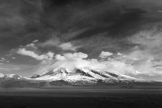 Black and white view of snow-capped Muztagh Peak crowned with a cap cloud, under a dramatic moody sky, with vast arid plains below, capturing the stark wilderness of the Pamir Plateau alone Kkh
