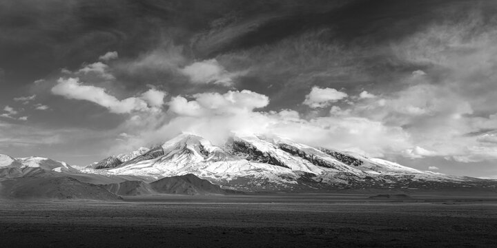 Black and white view of snow-capped Muztagh Peak crowned with a cap cloud, under a dramatic moody sky, with vast arid plains below, capturing the stark wilderness of the Pamir Plateau alone Kkh