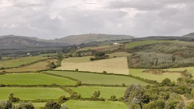 Rolling green hills and patchwork agricultural fields stretch across the rural Irish countryside on a cloudy day.