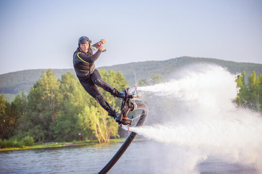 A flyboarder performs an impressive stunt above the shimmering water, propelled by powerful jets. The backdrop features lush green trees and rolling hills, highlighting a beautiful natural setting.