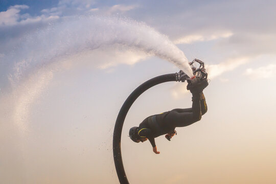 Athlete deftly maneuvers on a flyboard, soaring high above the water with a powerful jet of water propelling them up. The background highlights a beautiful sunset, casting warm hues across the sky.