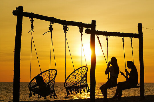 Two people sit on swings at a beach as the sun sets behind them. The sky shows warm colors while the sea reflects the light of the setting sun.