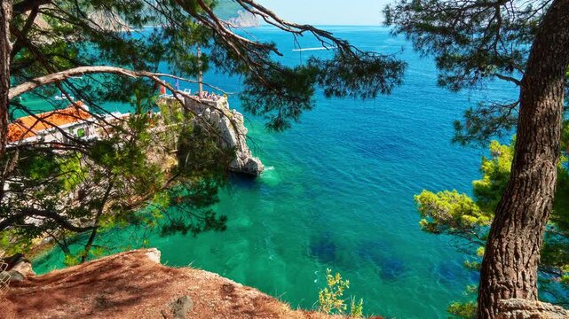 Beautiful view of the Adriatic coast in Petrovac, Montenegro, featuring a stone path, lush pine trees, and a tourist boat on turquoise water. Perfect for summer travel and Mediterranean vacation theme
